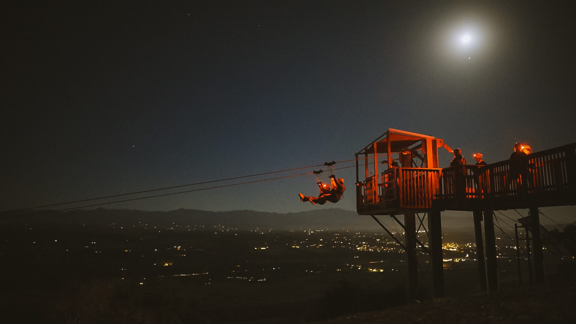Zipliner flying under a full moon with the valley lights below on the Moonlight Zipline Adventure