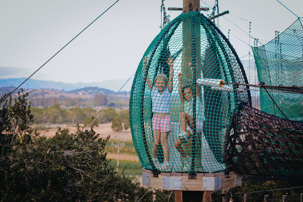 Two kids inside a suspended net pod of the Skynet Playground with the Santa Ynez Valley behind them