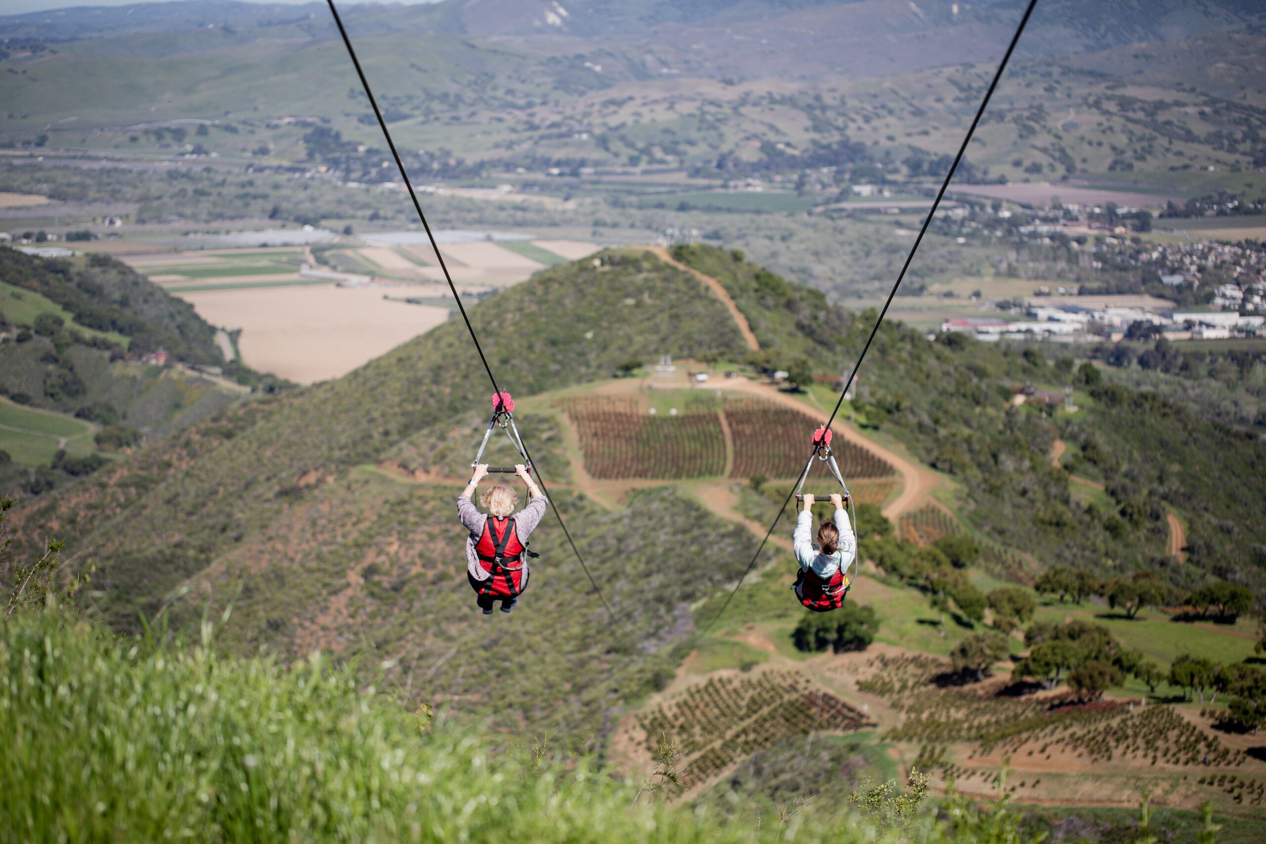 Two first-time zipliners flying side by side on the dual lines above the green hills of the Santa Ynez Valley at Highline Adventures
