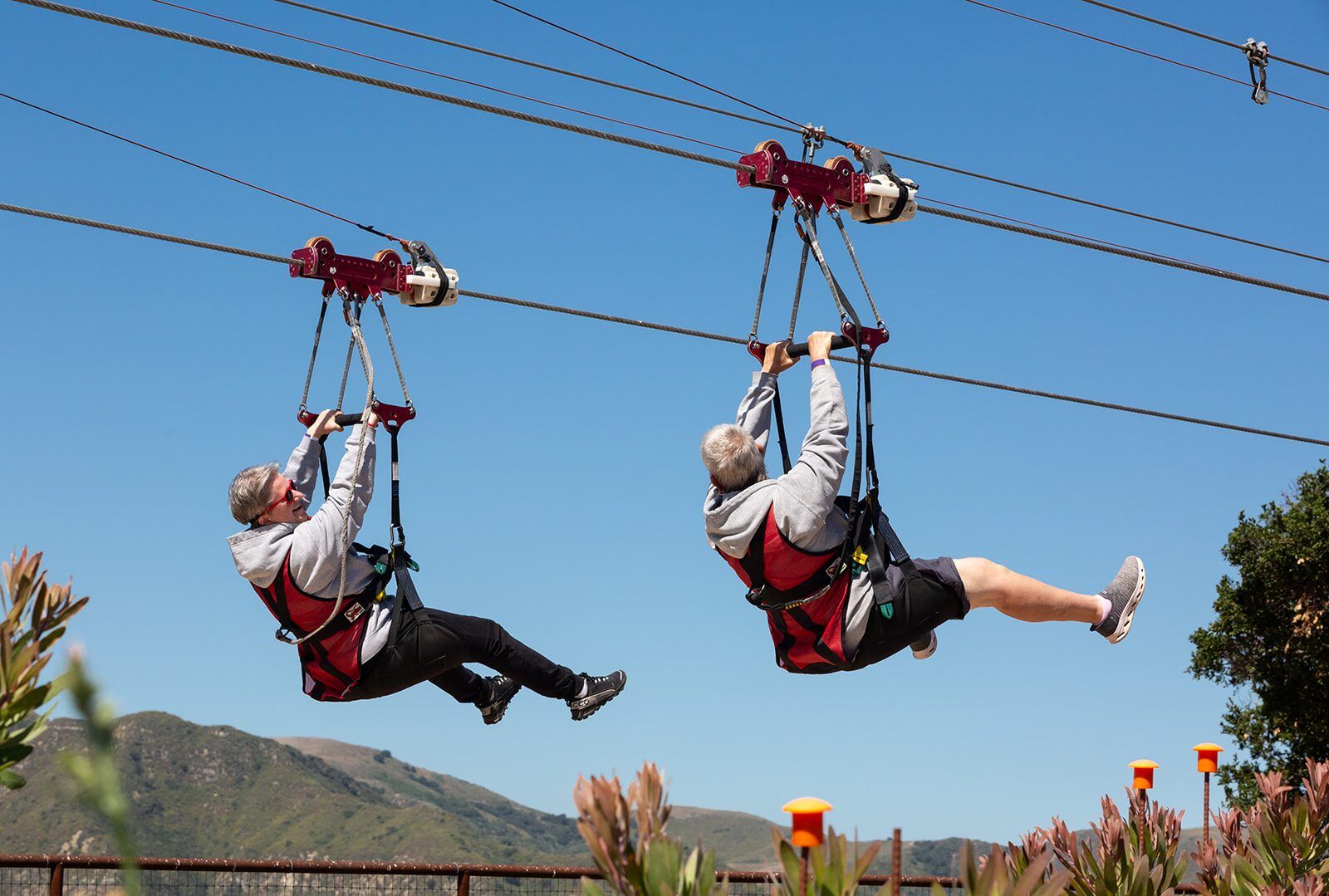 Two zipline riders side by side on parallel cables high above the Santa Ynez Valley hills.
