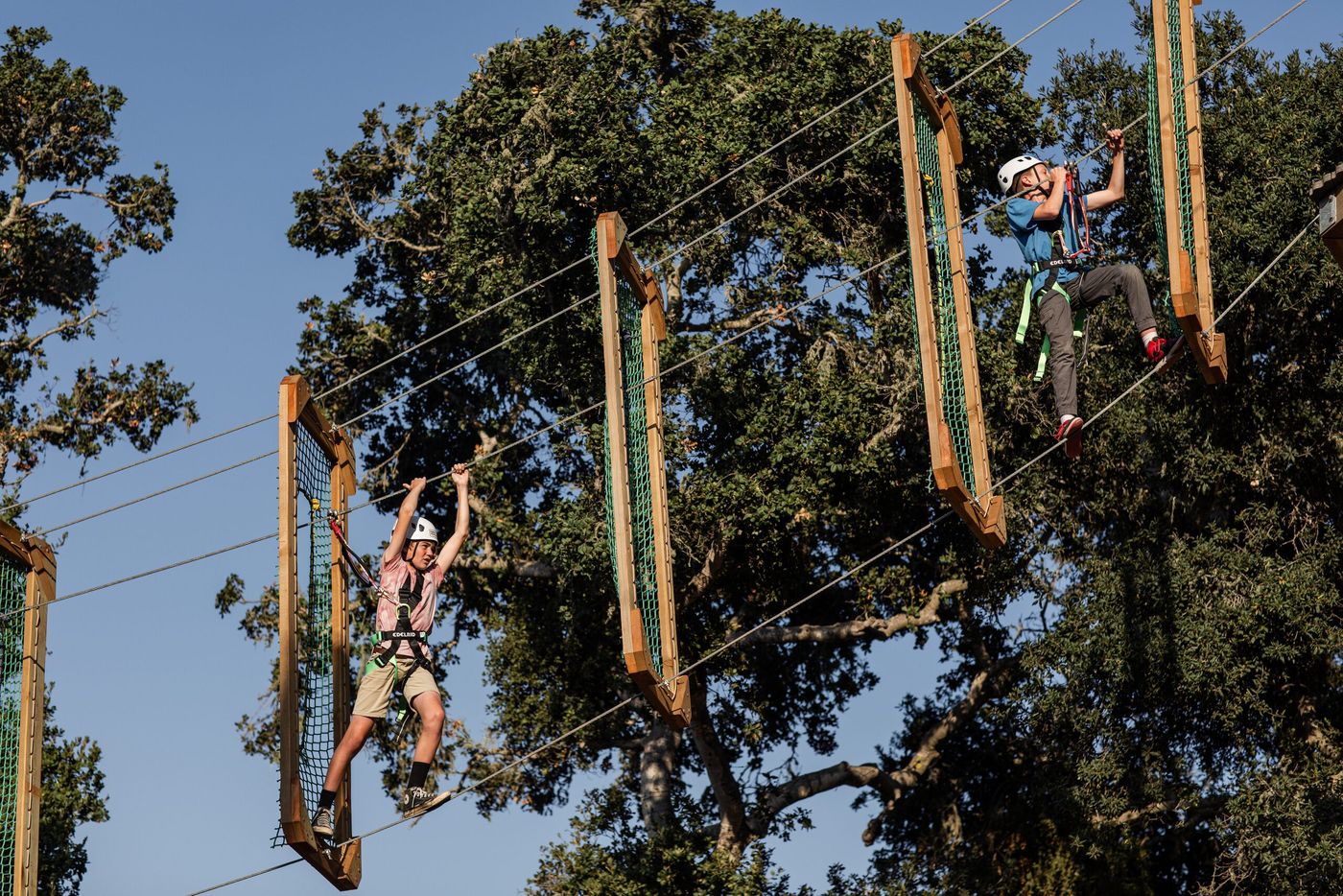 Two young climbers in helmets and harnesses crossing high elements of the treetop ropes course against a blue sky.