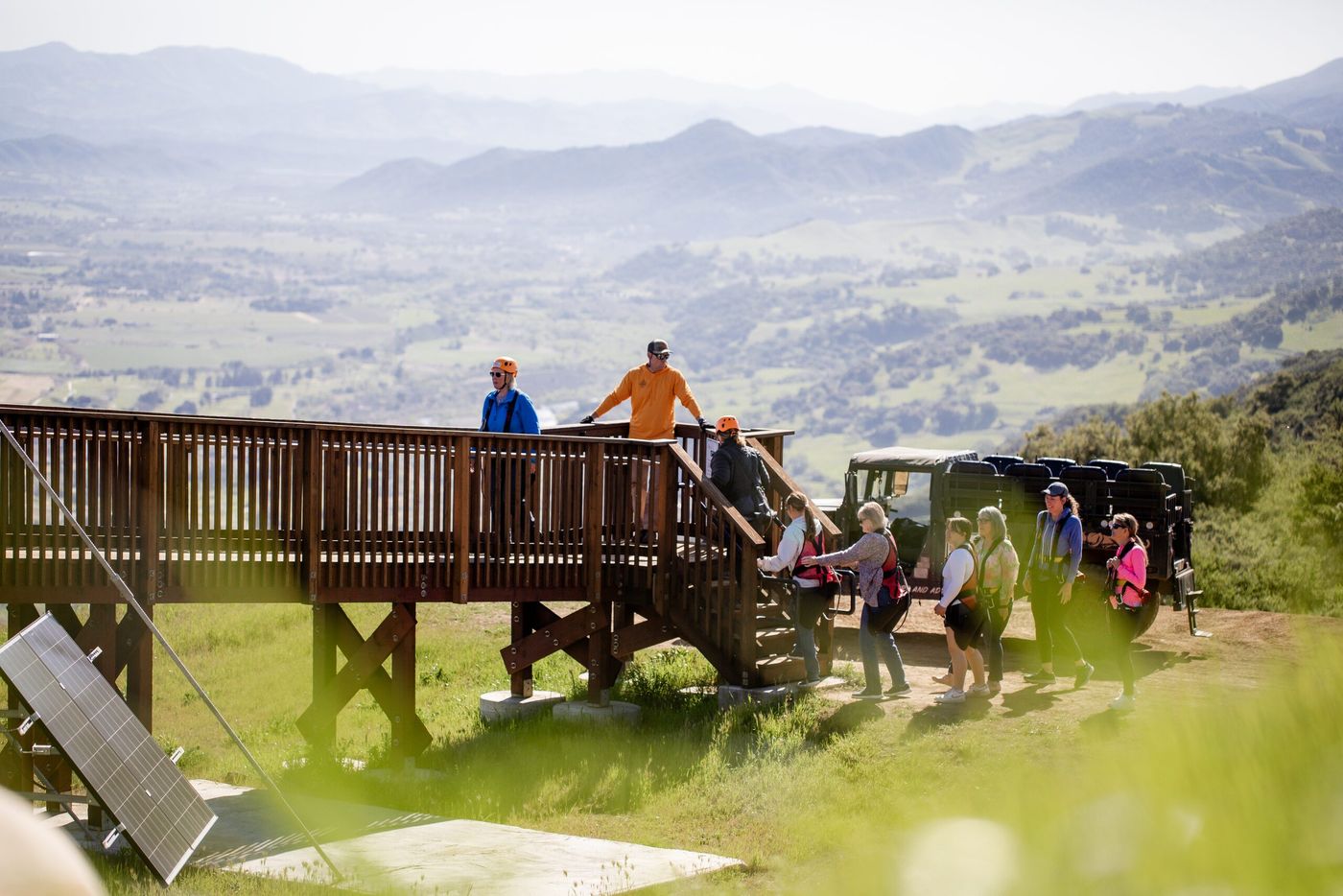 A group gathered on the wooden summit platform beside the 4x4 Humvee, with green hills and the valley below.