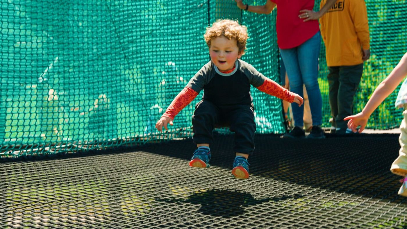 A small child mid-jump on the suspended net of Skynet Playground, with family members standing behind.