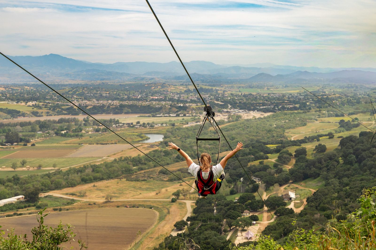 A zipline rider with arms spread wide flying over the green farmland of the Santa Ynez Valley in spring.