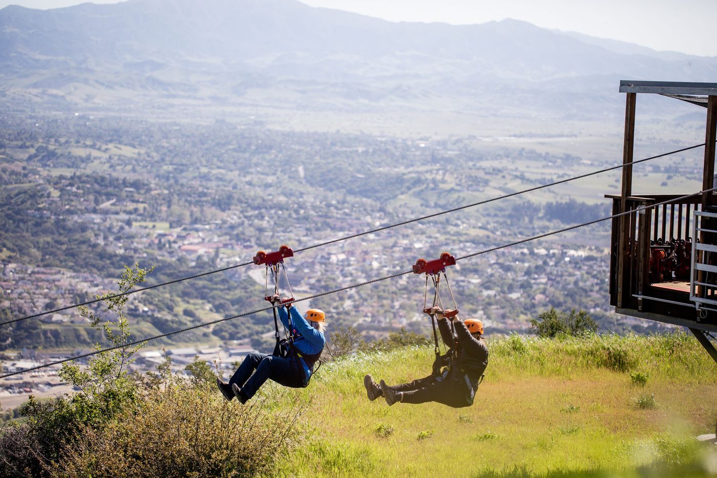 Two zipline riders side by side leaving the platform above the green hills of the Santa Ynez Valley with the town below.