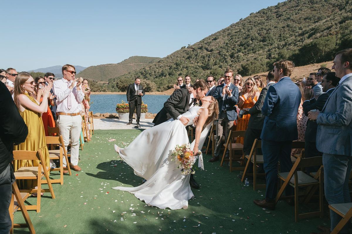 Tiffany and Andrew's first kiss on the lakeside lawn, guests standing on either side of the aisle.